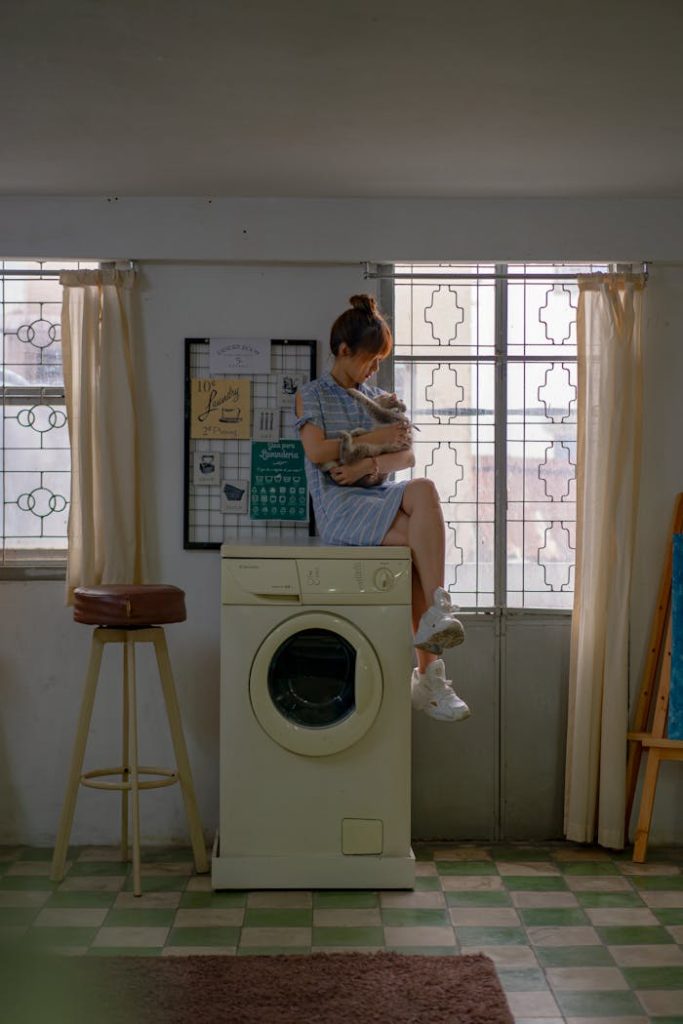 Woman in blue dress sitting on washing machine. Indoor laundry room setup with window light.